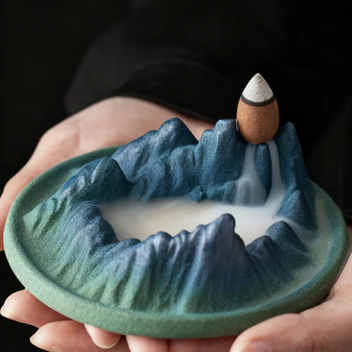 Ceramic mountain landscape with a incense waterfall held in hands against a dark background