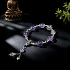 Purple beaded bracelet with silver charms on a dark surface, with a small statue and plant in the background.