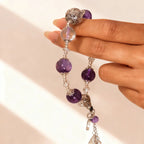 Hand holding a beaded necklace with purple beads and silver accents against a light background